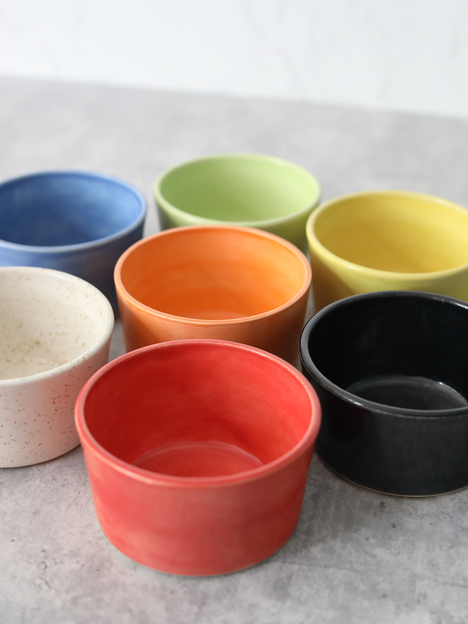Seven handmade ceramic side bowls arranged in a cluster on a grey marble surface, viewed from a slight angle to show the interiors, in blue, spring green, yellow, mandarin orange, speckled white, cherry red, and black glazes