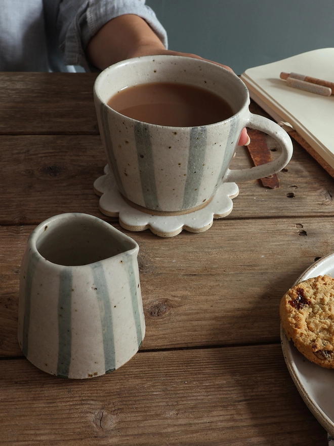 Handmade Ceramic Stoneware Small Jug in Blue and White Stripe sat on a wooden surface in front of a mug in the same style filled with coffee. A female can be seen sitting behind the table, holding the mug of coffee. The mug sits on top of a handmade flower coaster. A notebook and pen, and a plate of cookies also sit on the table. 