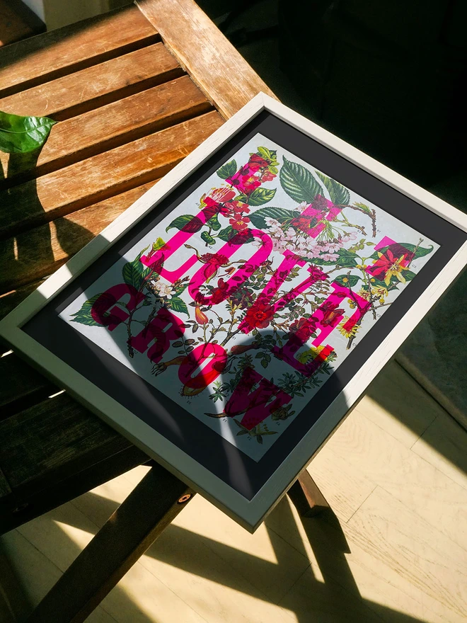 A framed limited edition artwork rests on a wooden surface, partially lit by sunlight. The letterpress print features a mix of bright pink and green leaves with the phrase "LET LOVE GROW" in bold, colorful letters rendered in fluorescent ink.