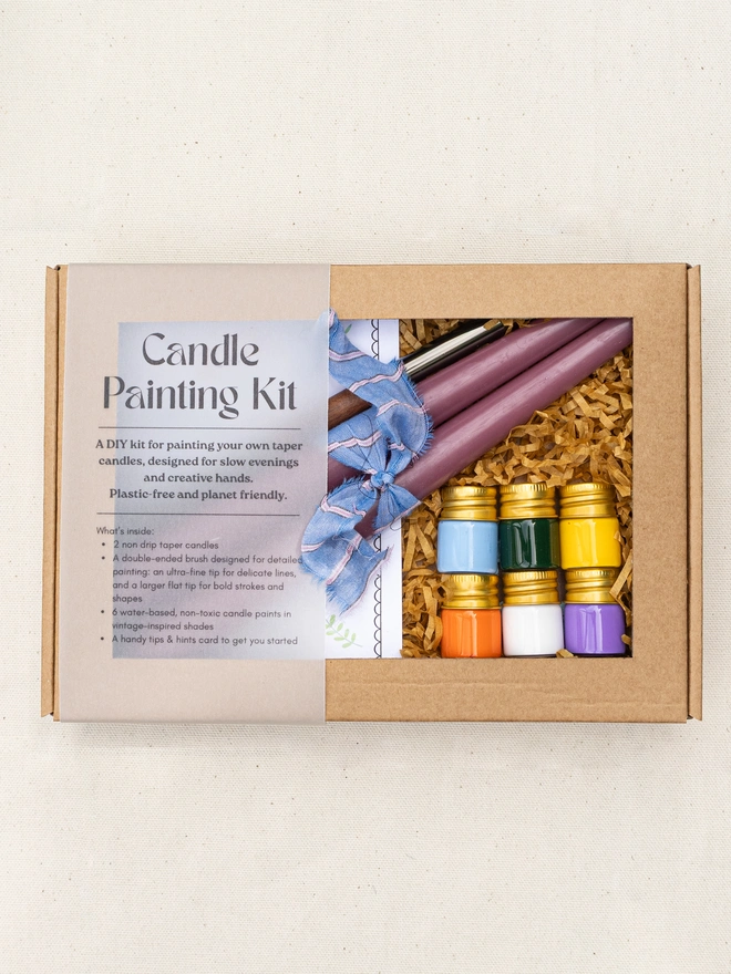 Candle painting kit gift box containing two mauve taper candles, six pots of colourful candle paint, a double-ended paintbrush and an illustrated instruction booklet in a kraft presentation box.