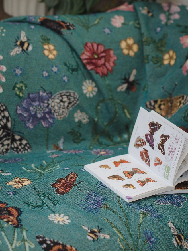 'Pollination Bloom' Recycled Cotton Blanket by Arcana seen on a sofa with a book of butterflies.