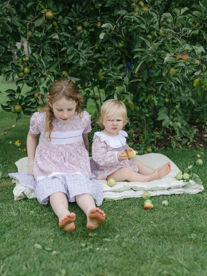 A litttle girl in a romper sits with a  bigger girl in a floral and check dress under an apple tree