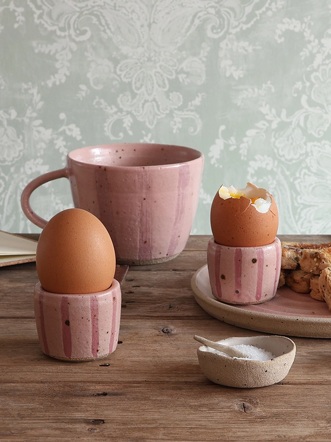 Two pink-striped ceramic egg cups holding boiled eggs sit on a wooden table alongside a matching plate of buttered toast and a mug. 