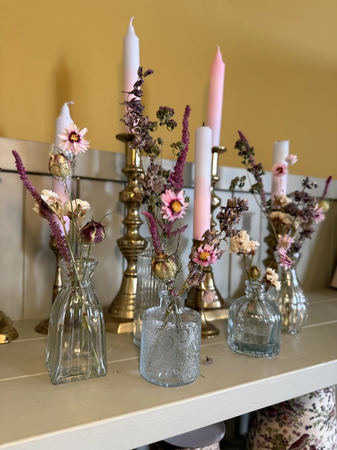a selection of dried flowers in glass bud vases on a shelf with candles in brass candlesticks