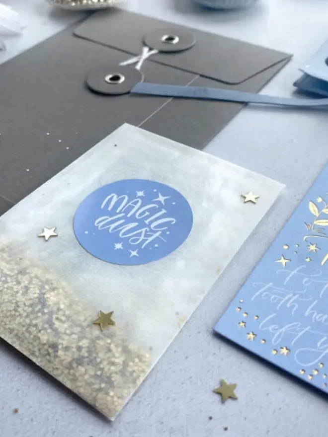A glassine packet of gold glitter with a blue "Magic Dust" sticker and gold confetti stars. A grey string-tie envelope and blue notecard sit beside it.