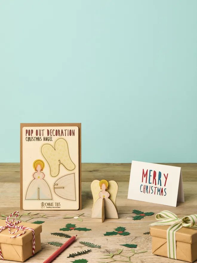 Pop out laser-cut Angel Christmas decoration and Merry Christmas card and brown kraft envelope on top of a wooden desk in front of a sky blue coloured background