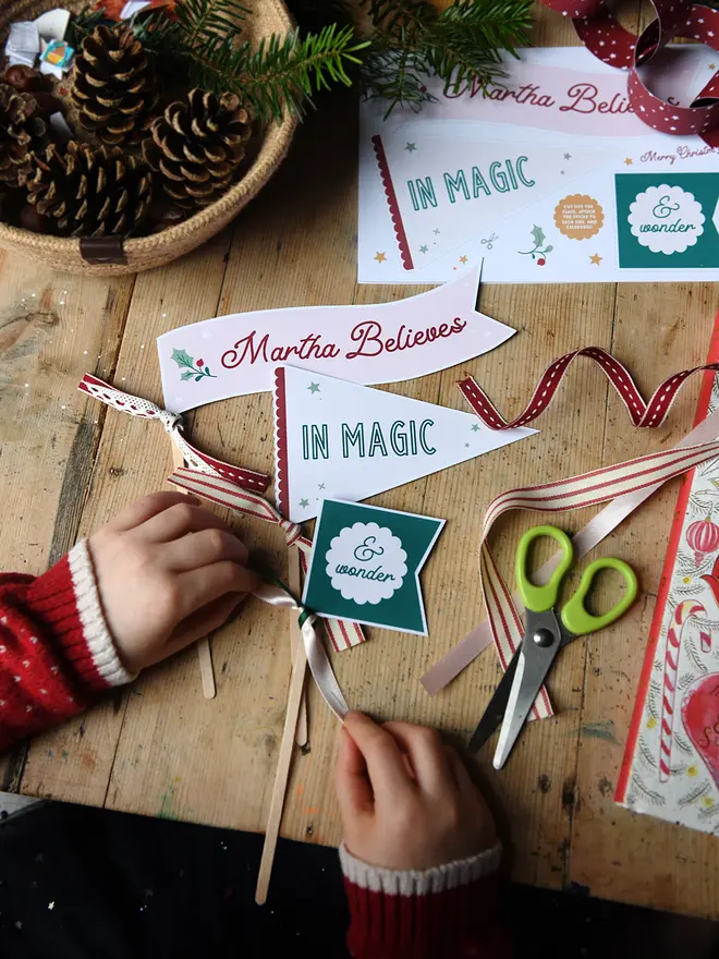 A young child is crafting Christmas flags with messages of magic and wonder printed on them. They are laid on a wooden desk with various traditional Christmas items around them.
