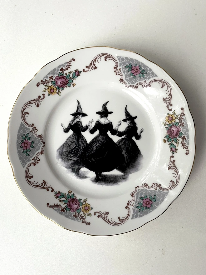 a coven of 3 witches dance hand in hand, the image is victorian in black and white and is printed directly onto an ornate vintage plate