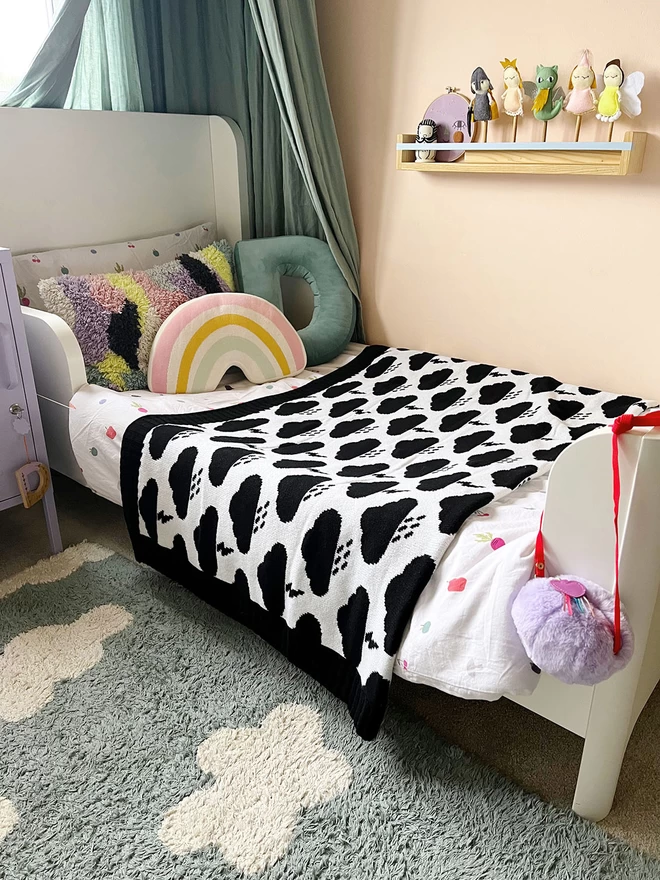 A warm and inviting childrens room showing a white extendable toddler bed with a duck egg blue canopy hanging above the head. Across the middle of the bed is a folded white blanket with black storm clouds with raindrops and lightning bolts on. A rainbow cushion sits by the pillow and toys adorn a shelf on the wall.