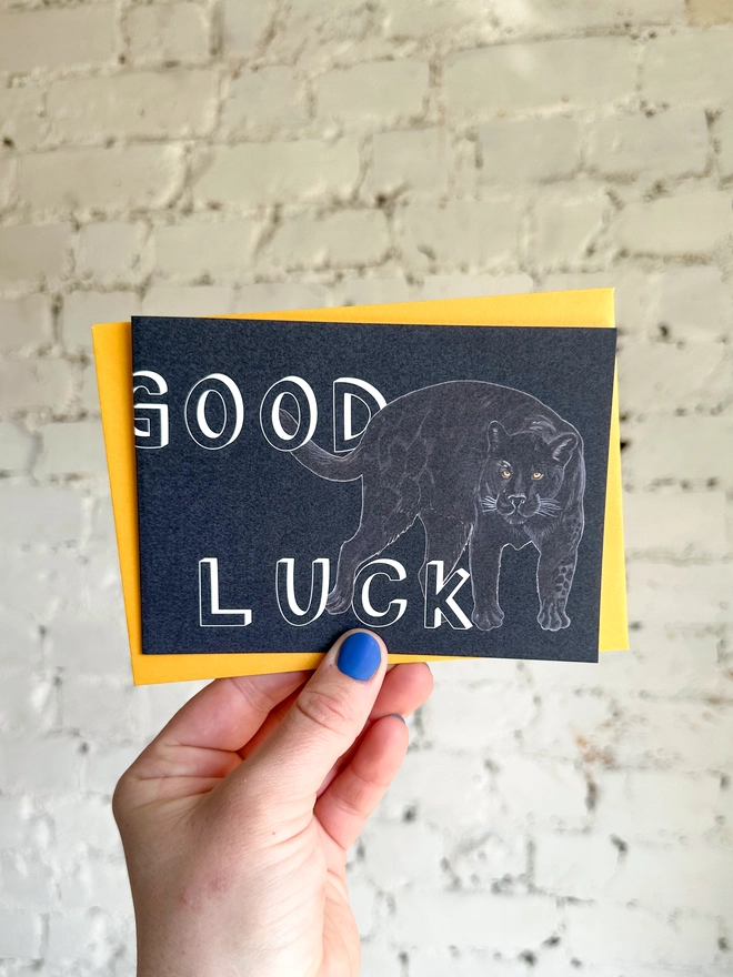 a dark grey greetings card featuring an illustration of a black panther with the text “good luck” with a deep yellow envelope
