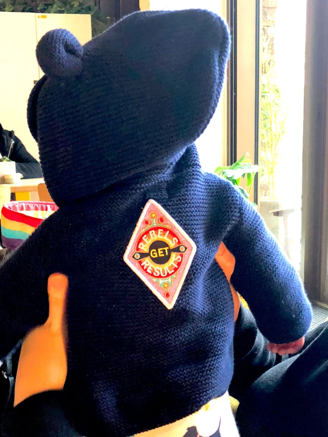 A toddler is dressed in a dark blue, hooded top with the patch on the back. The patch is a diamond shape embroidered patch with “Rebels Get Results” at the centre. The design features pink, white, red, yellow and black colours and has a white border.