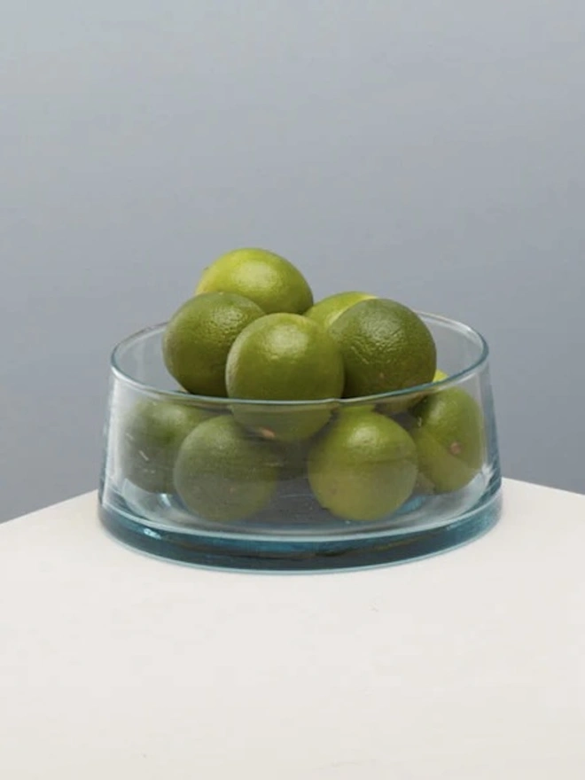 Low Glass Dish, a small glass dish sitting on a white counter top against a baby blue backdrop. It is filled by some limes. 