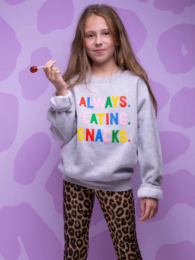 A young girl wearing a grey sweatshirt with multi coloured Always Eating Snacks slogan.
