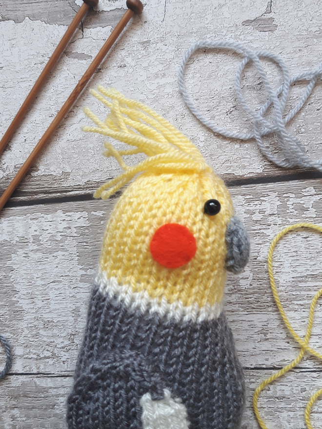 a knitted cockatiel lying on a wooden table with knitting needles and yarn