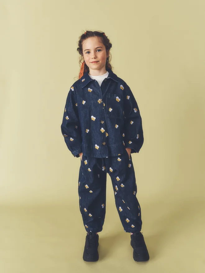 Child stands with hands in pockets against a pale yellow backdrop, wearing a matching Studio Koter dark denim set with Denim Petal embroidery: button-front overshirt and relaxed trousers.