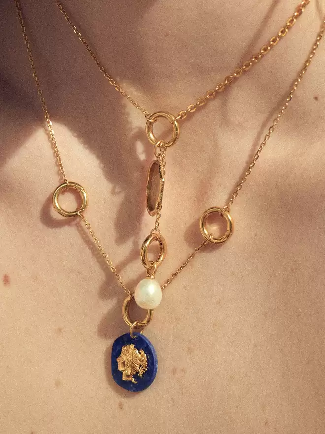 woman wearing two gold necklaces featuring a pearl charm and a lapis lazuli Aphrodite pendant.