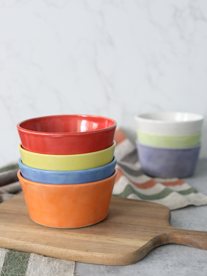 A stack of four glazed ceramic breakfast bowls in orange, blue, yellow, and red, resting on a wooden chopping board, with a stack of further pasta bowls partially visible in the background.
