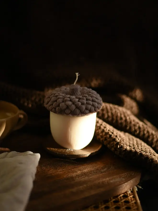 The Acorn Candle. An acorn candle with a dark brown cap and cream base on a small wooden coaster, styled with a chunky knit throw and a ceramic cup. The cosy seasonal candle makes a thoughtful autumn gift for nature lovers.