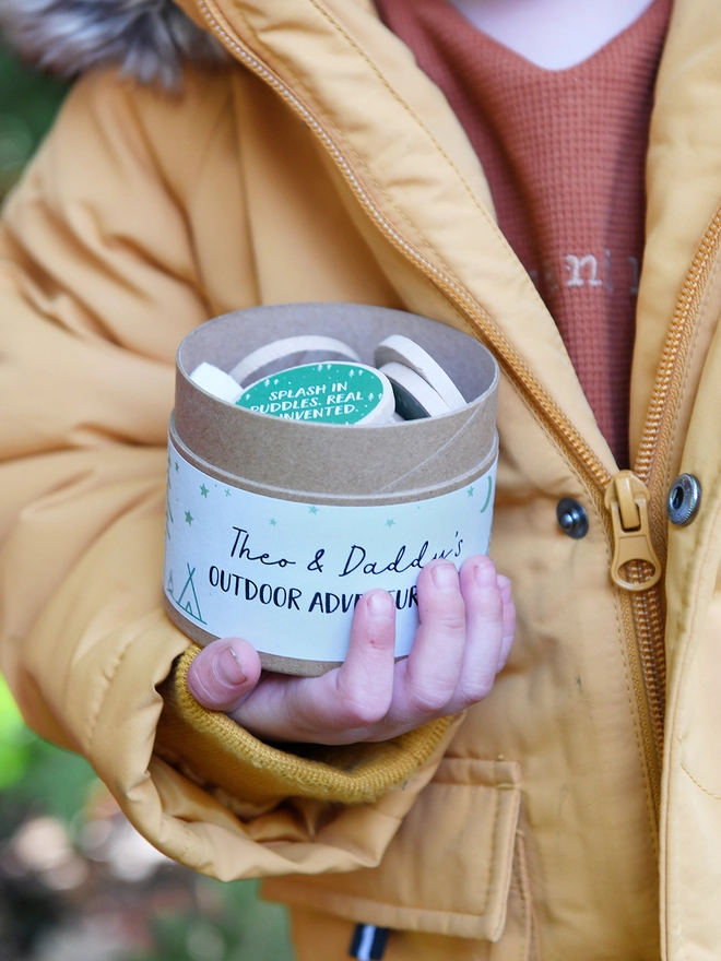 A young child wearing a mustard yellow coat holds a cardboard jar with several wooden tokens poking out of the top. A label on the outside reads Outdoor Adventure Ideas.