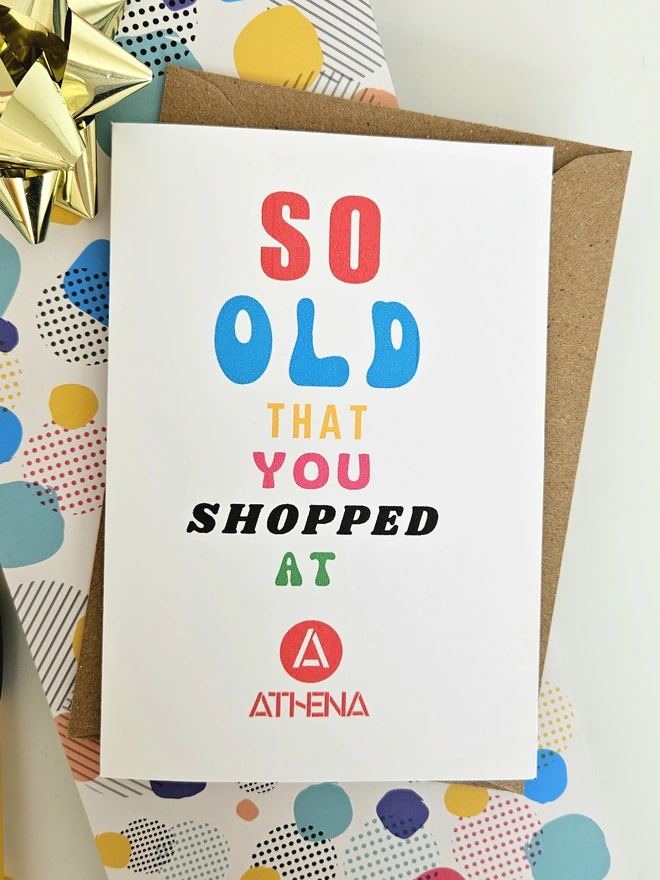 A photograph of a humorous A6 greeting card illustrated using brightly coloured text and the retro shop logo. The text says so old you shopped at Athena.