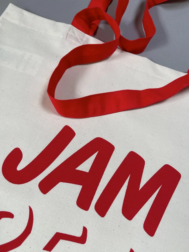  Close up of a white, natural coloured tote bag laid down with the word JAM written on top of CREAM, screenprinted in red ink. The handles are red to match the printing.