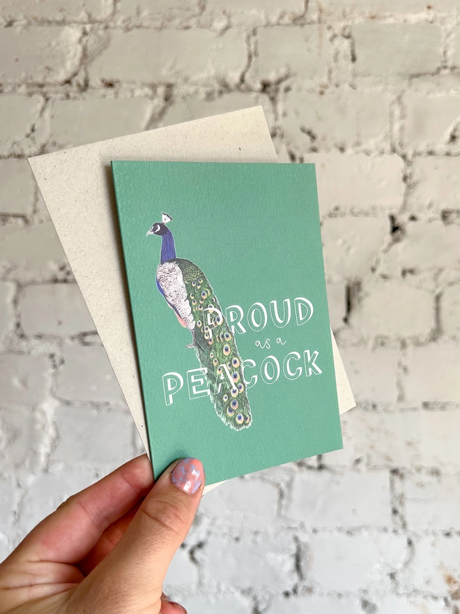 a deep green greetings card featuring an illustration of a peacock with the text “proud of a peacock”