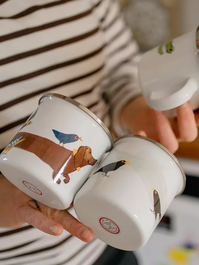 Hands hold three shiny enamel mugs illustrated with birds and animals, against the background of a striped top