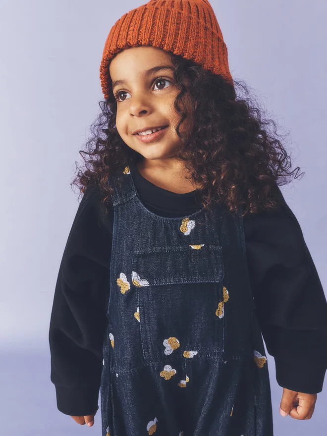 Child smiles holding a daisy against a lilac backdrop, wearing Studio Koter Kai dungarees in Denim Petal, dark denim with floral embroidery, over a black sweatshirt with yellow socks and black sandals.