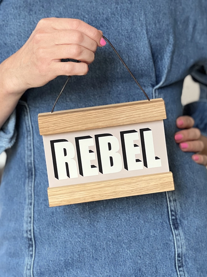 A REBEL print with a pale pink background, framed in a oak hanger, held by a woman wearing pink nail varnish 