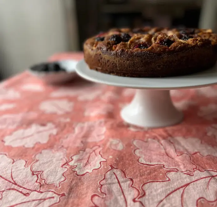 Grace Oak Trees Pink Linen Tablecloth. Light Pink and white tablecloth with oak patterns, styled on an outdoor dining table with plates, glassware, and white floral arrangements.