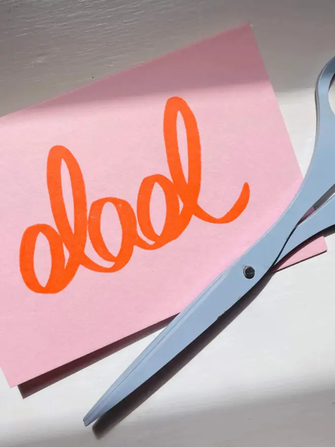 'Dad' hand printed in red on a pink card, displayed on a table, next to scissors. 