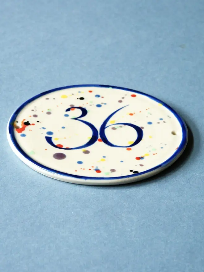 personalised hand-thrown ceramic house plaque. plaque features rainbow coloured dots and the number 36, photographed against a blue background.