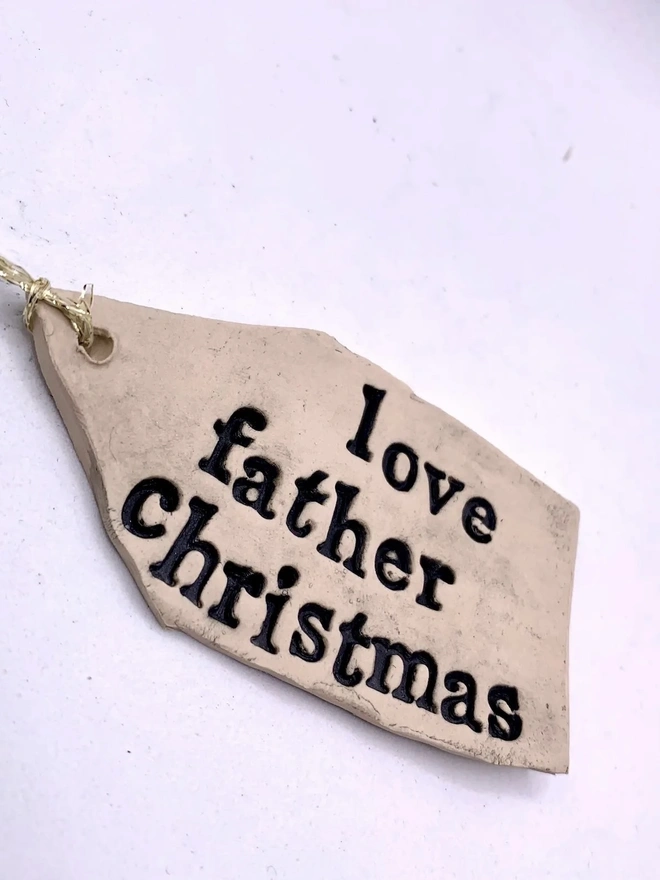 'Love Father Christmas' Handmade Ceramic Gift Tags, a ceramic gift tag attached sitting on a ale purple surface. 