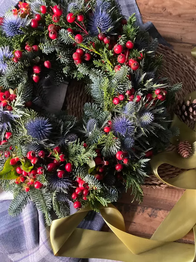 A Christmas Berry and  Fresh Eryngium Thistle Wreath sits on a wooden table, a pale blue tartan cloth sits alongside,  a green thick satin ribbon  is attached to the wreath.