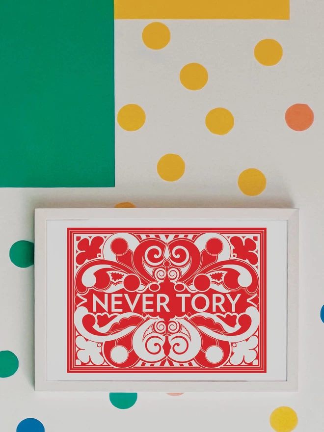 A bold, landscape red and white symmetrical illustration with Never Tory written in white at the centre. The picture is hanging in a white frame on a white wall, with yellow, orange, green and blue spots and a green and yellow rectangle painted in the top left hand corner.
