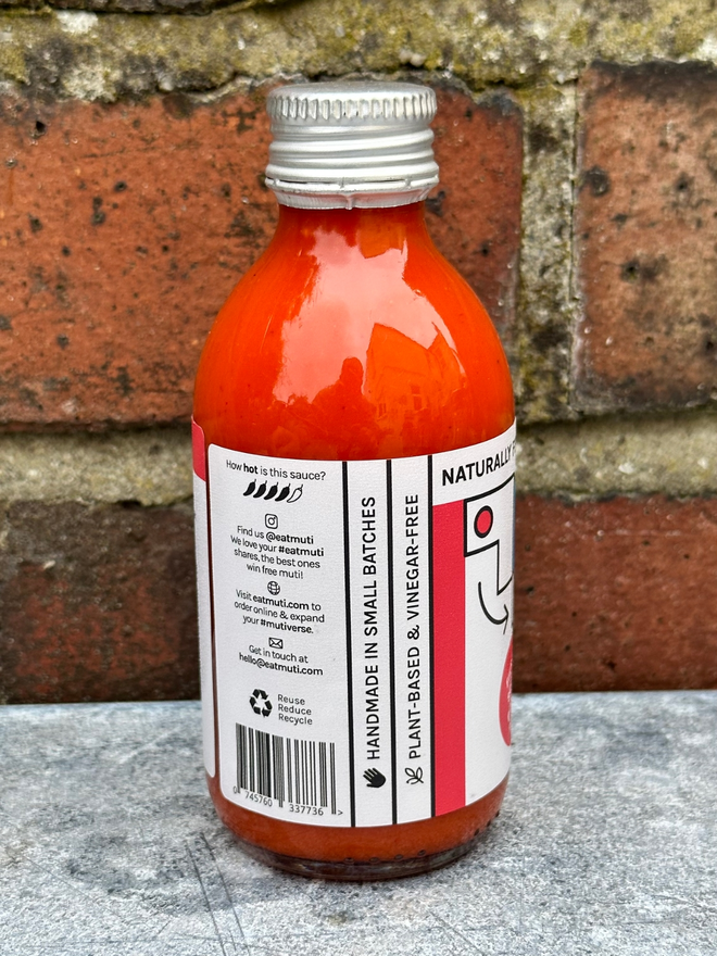 Picture Of Muti Double Trouble Bottle Facing Left And On A Steel Topped Table Against a Red Brick Wall.
