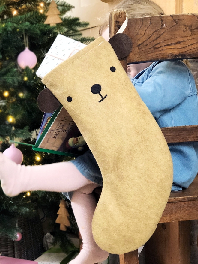 A handmade felt bear stocking hangs on a wooden chair, where a little girl sits, in front of a Christmas Tree.