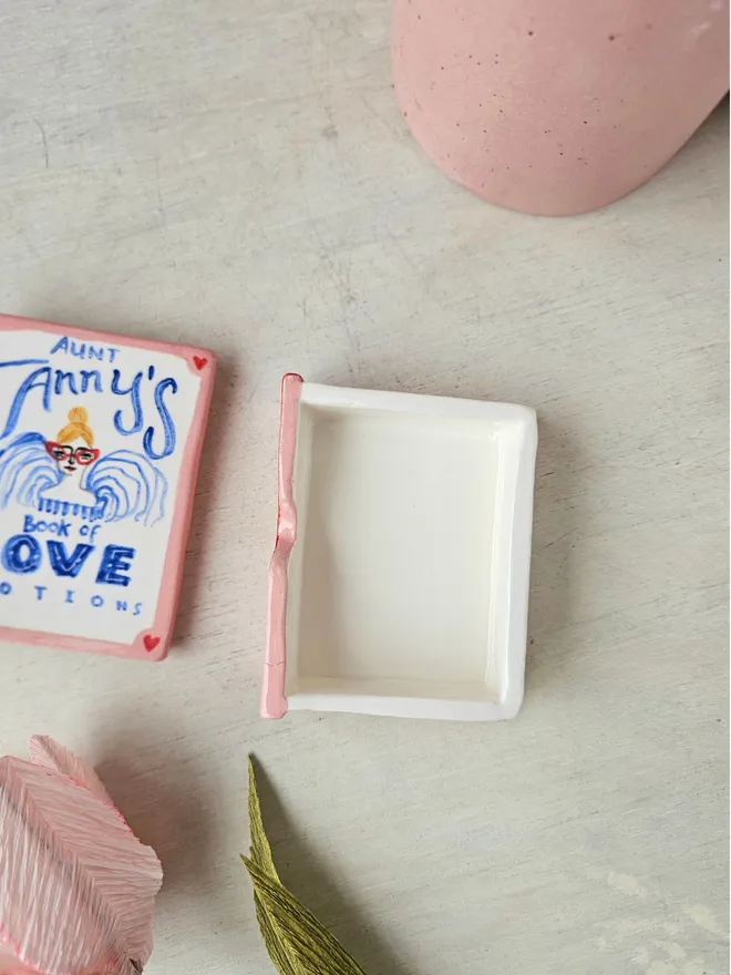  A charming ceramic trinket box designed to look like a vintage book, painted in shades of pink and red. The cover features the title "Aunt Fanny’s Book of Love Potions" in a playful, illustrated style. With its book-like shape and whimsical design, this decorative piece adds a touch of romance and mystery while offering a secret spot to store small treasures.