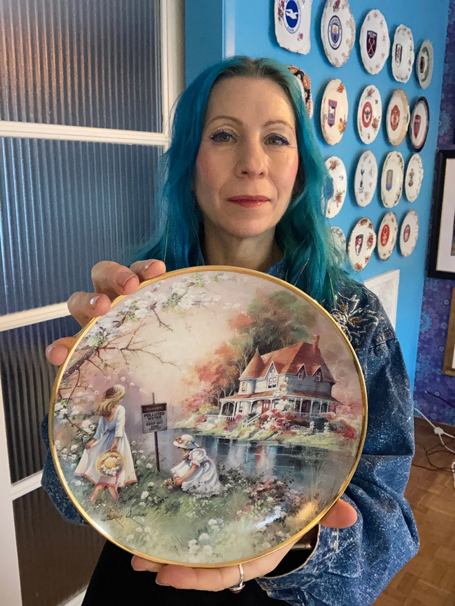 Lucy holding the Beautiful decorative 8” plate featuring a watercolour style image of two young girls playing by a river. On the other side of the river is a beautiful white house. The scene is idyllic, yet in the river we see a warning sign which says ‘Danger! Polluted Water’...