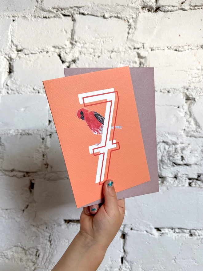 bright orange card with a big white number 7 and an illustrated madagascan fody bird with a purple envelope