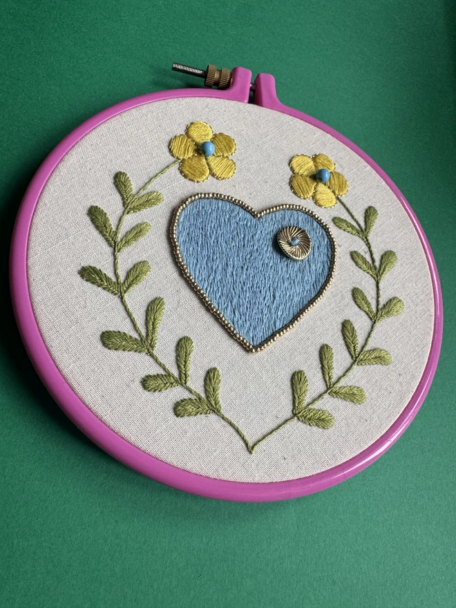 A pink embroidery hoop containing a stitched blue heart surrounded by two yellow flowers on long green stalks 