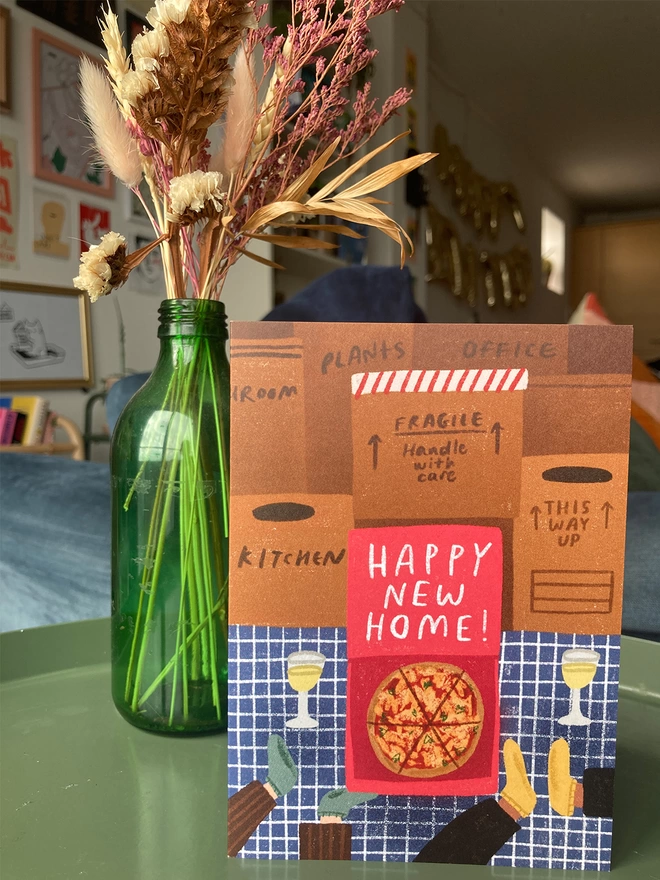 Greetings card with 'Happy New Home' message written on a takeaway box, surrounded by moving boxes. Placed on a table with flowers in the background.