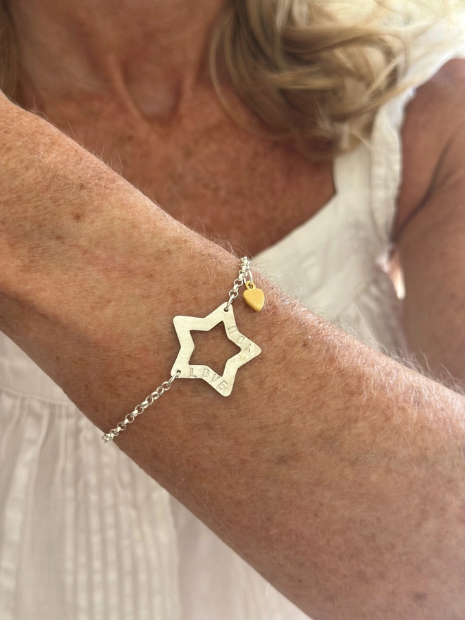 model wears a personalised sterling silver open star charm on a silver belcher chain bracelet with additional tiny heart charm in gold