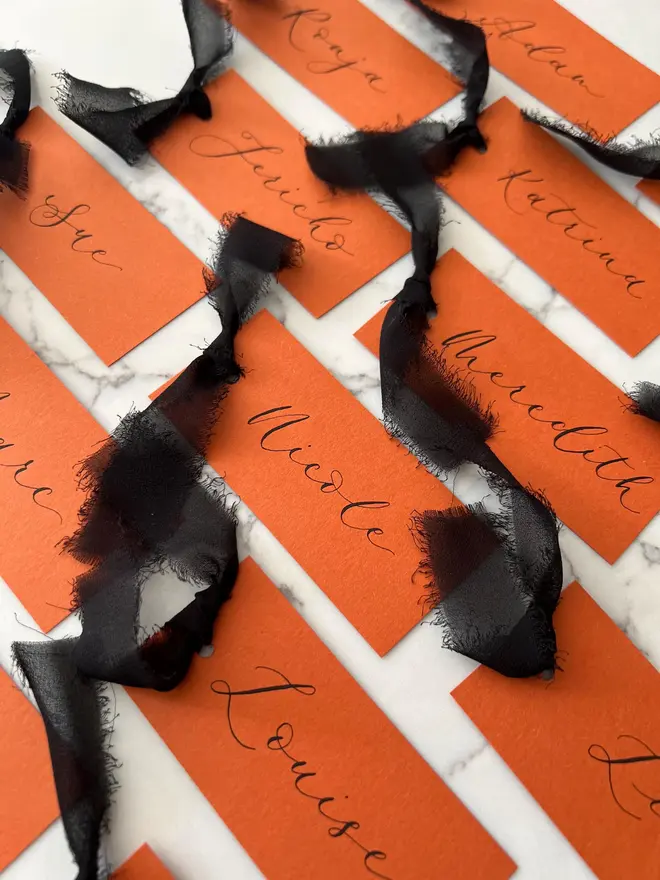 Personalised Calligraphy Rust Orange Place Card, orange place cards with black chiffon ribbons grouped together on a white surface. 