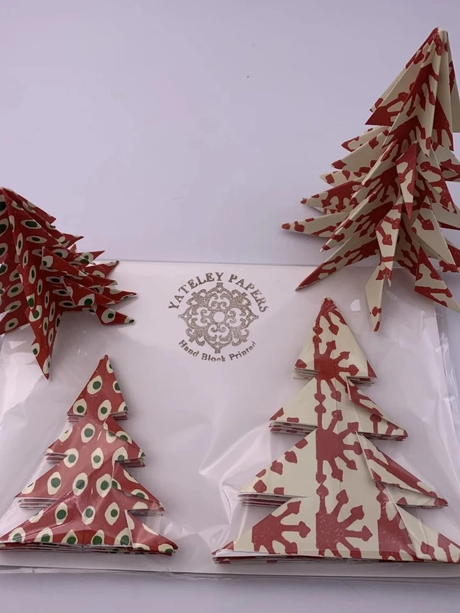 Origami Christmas Tree Kits (Set Of 2). Two folded paper Christmas trees in a clear cellophane pack with a Yateley Papers hand block printed backing card. One tree is red with cream and green spots and the other is cream with a red sunburst print, ready to give as a festive gift or decoration.