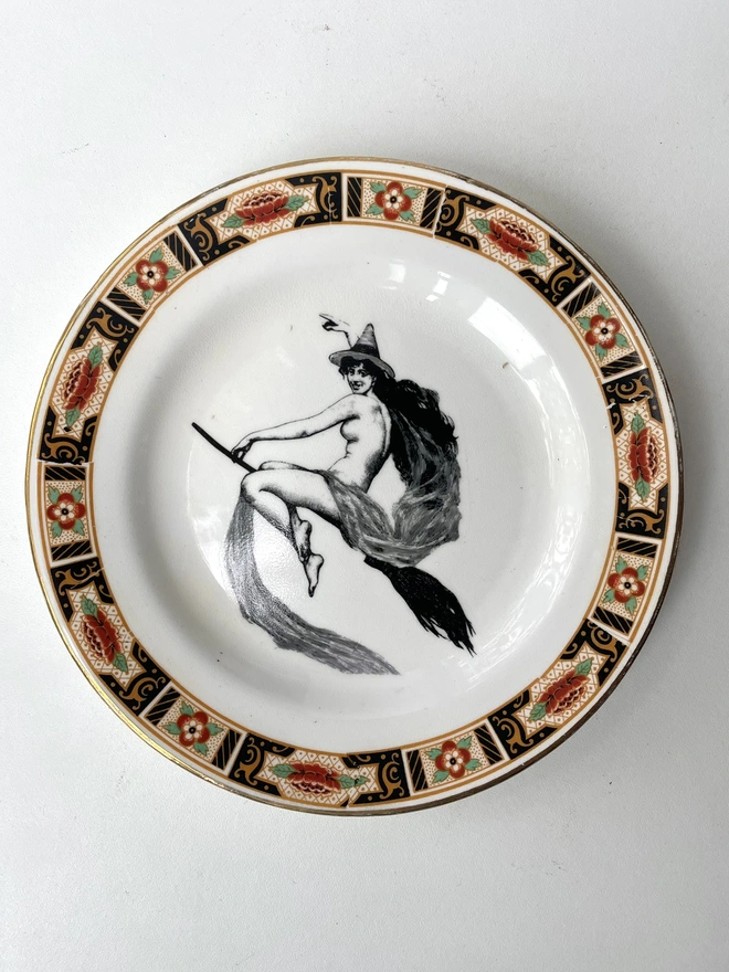 vintage plate with an ornate border, with a printed vintage illustration of a witch on a broomstick in the middle 