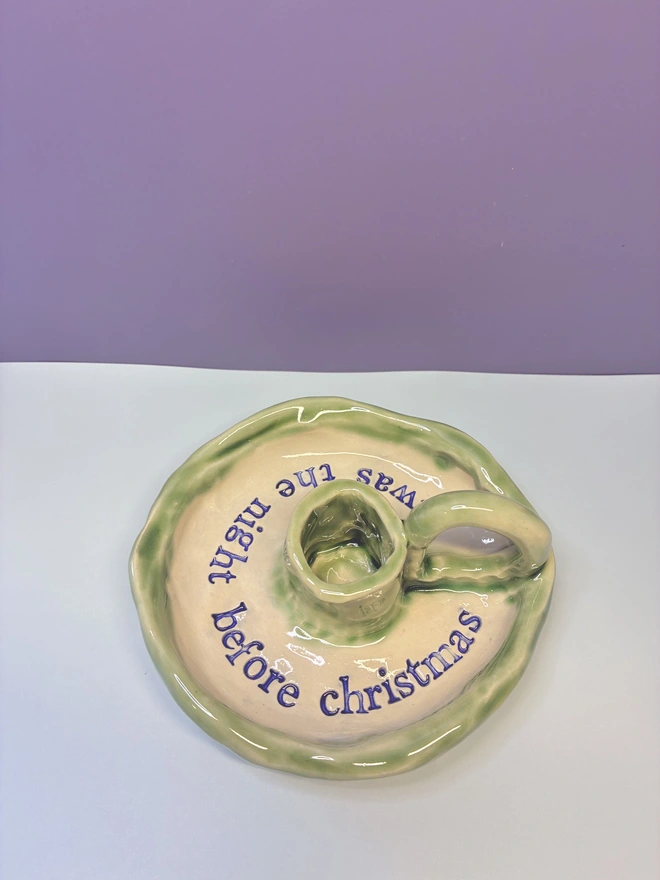 Handmade ceramic Christmas taper candlestick holder with a soft green glazed edge and the words “‘Twas the night before Christmas” in dark blue lettering, arranged around the candle holder opening. Photographed against a pale background.