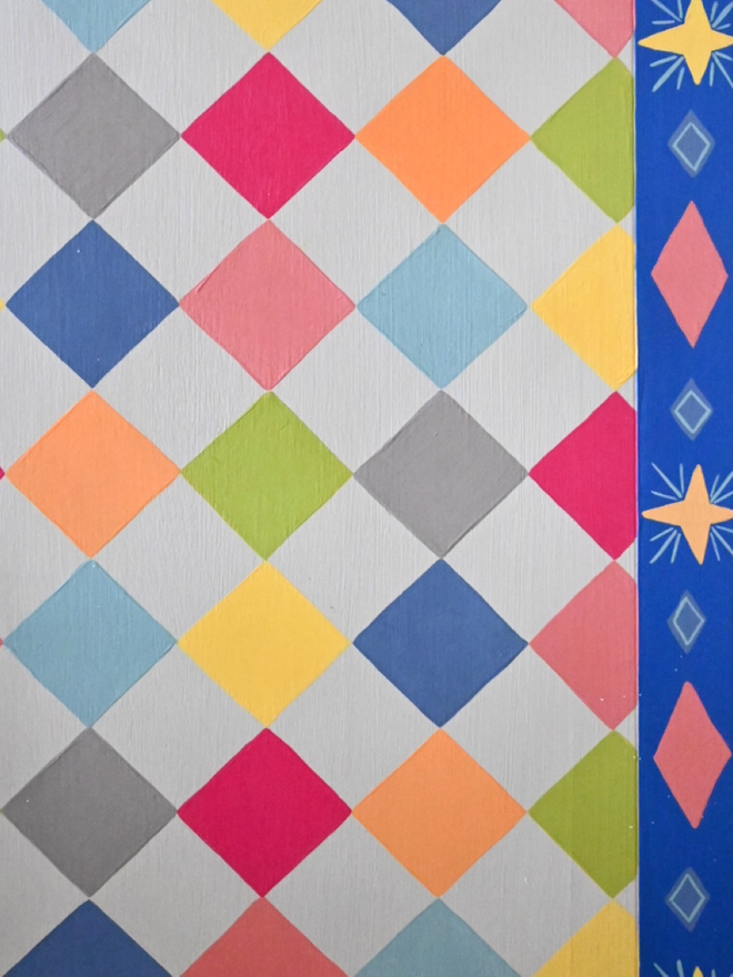 A close-up view of a repeating harlequin or diamond pattern. The diamonds are various bright pastel colors including pink, green, yellow, orange, and blue, set against a light gray or off-white background. A thick border of royal blue runs vertically along both sides, featuring a repeating design of smaller pink diamonds outlined in white, alternating with yellow starbursts or asterisk-like shapes. The image appears to be of painted wood or a similar textured surface.