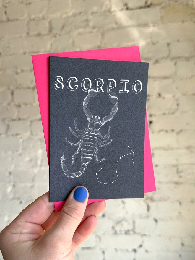 a greetings card featuring a greyscale illustration of a zodiac style scorpion with the text “SCORPIO” with a neon pink envelope