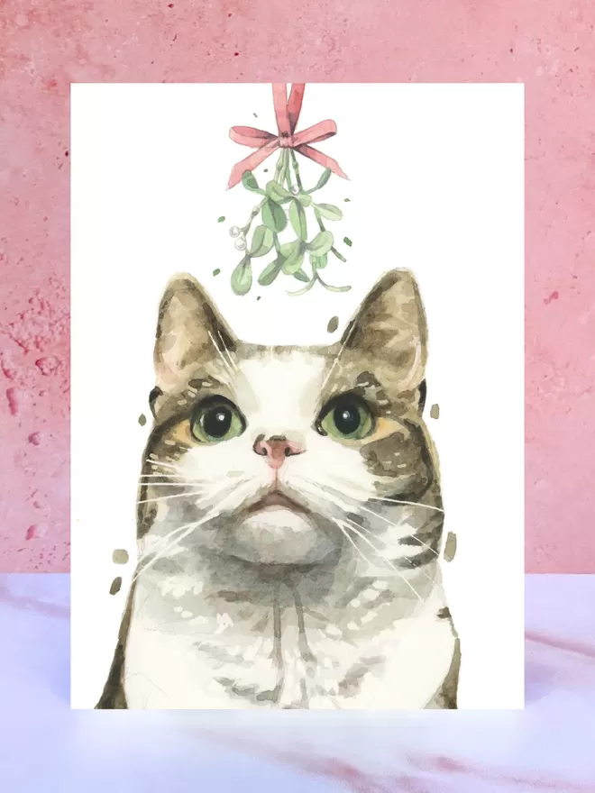 A Christmas card featuring a hand painted design of a Tabby and White Cat, stood upright on a marble surface.